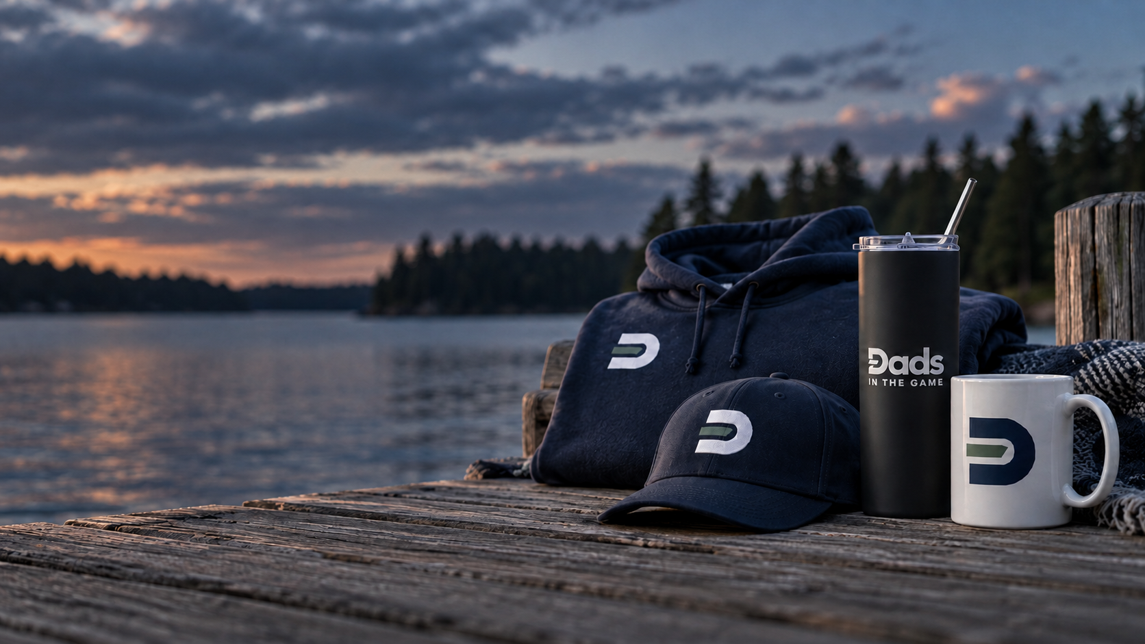 Dads in the Game hoodie, hat, tumbler, and mug on a dock at dusk